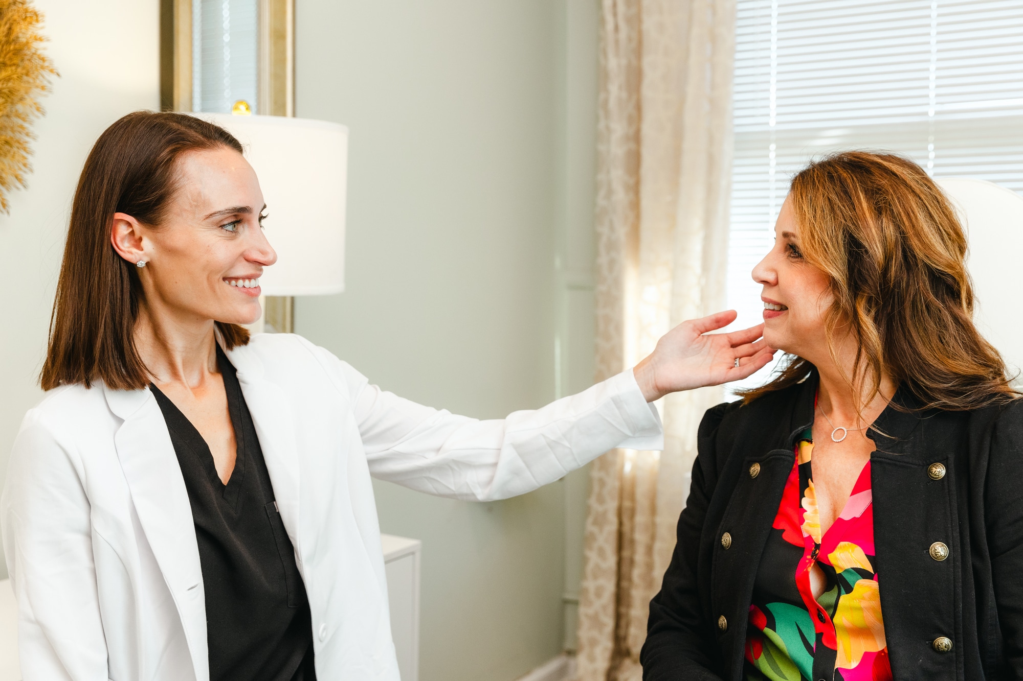 A provider sits alongside a patient at DeLozier Aesthetics, working together to tailor facials in Brentwood, TN, to meet the patient's needs. Both women are smiling, the provider using her hand to assess the patient's skin concerns.